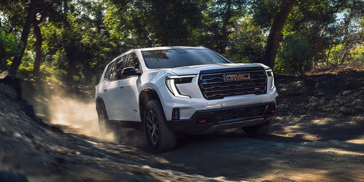 Exterior View of the 2026 GMC Acadia Mid-Size SUV Driving on a Dirt Path Surrounded by Trees