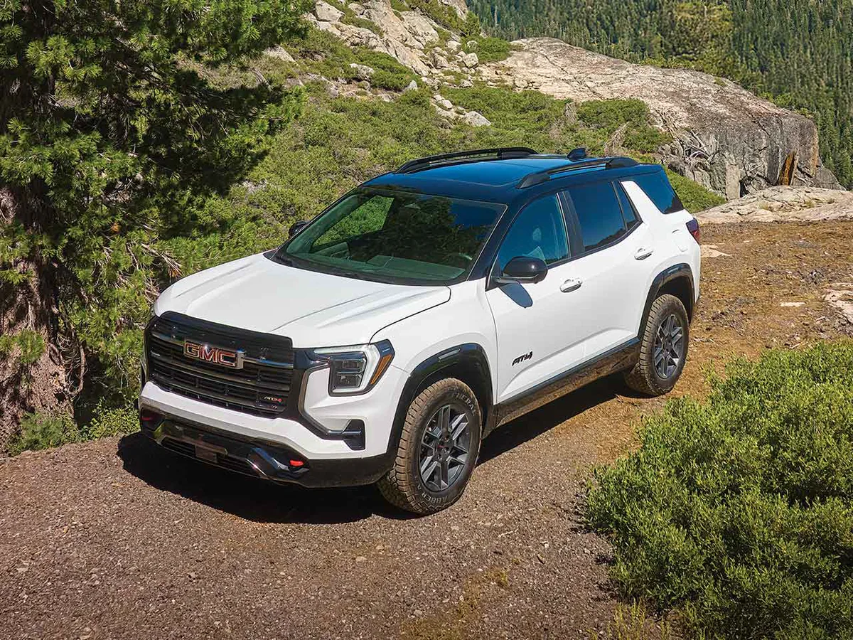 A White GMC AT4 SUV Parked on a Dirt Trail Beside Green Trees And Rocky Terrain Under a Clear Blue Sky